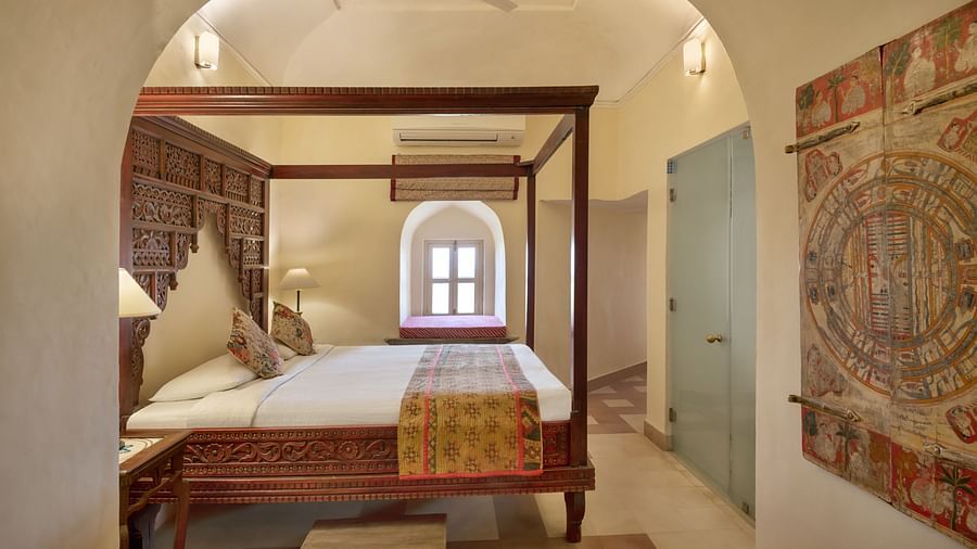 A room with a four-poster bed, a window with an arched top, and a framed artwork, seen through an arched doorway in Tijara Fort-Palace - 19th Century, Alwar.