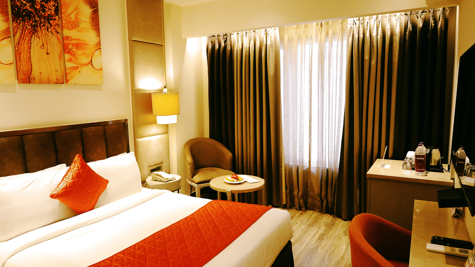 A room at Hotel Saket 27 features a king size bed, upholstered headboard, bedside table, paintings on the wall, desk, chair, television, table lamp and large curtained windows.