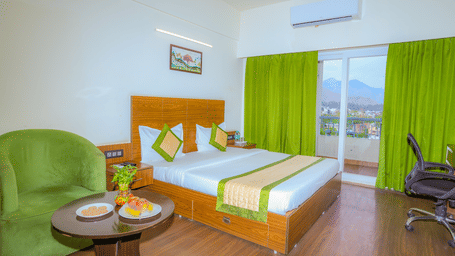 A king size bed in the Premium Room with Balcony at Livbox, Dehradun, which also features wooden flooring, curtained windows opening to the views of the surrounding, and a seating area.