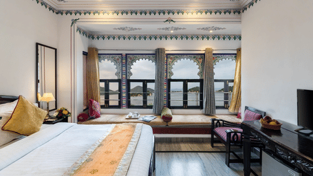 A side view of the bedroom featuring a spacious bed, a wooden table under the wall-mounted television, and bay window seating with curtains pulled to the side at Panna Vilas Palace