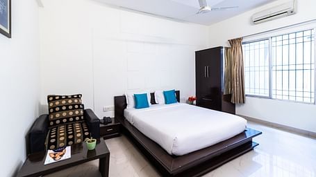 A room with a bed, an armchair, a wardrobe, and a large window at Sanctum Suites Domlur, Bangalore.