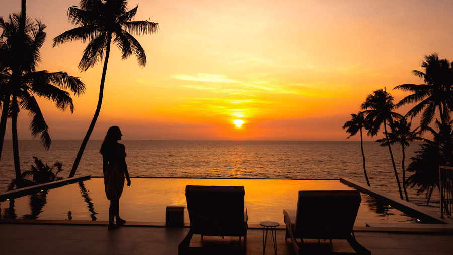 Silhouettes of a person standing and two lounge chairs beside an infinity pool at sunset at SUKAVI, surrounded by palm trees and overlooking the horizon.
