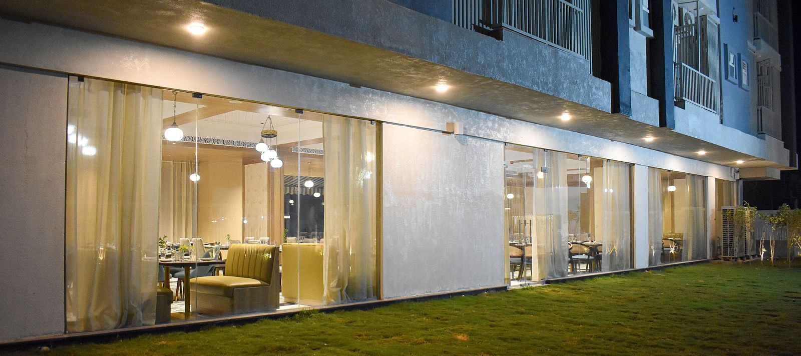 Exterior view of a building with a glass-walled restaurant, sheer curtains, hanging lights, and a grassy area in front - Urban Suites by BluSalzz, Jaipur