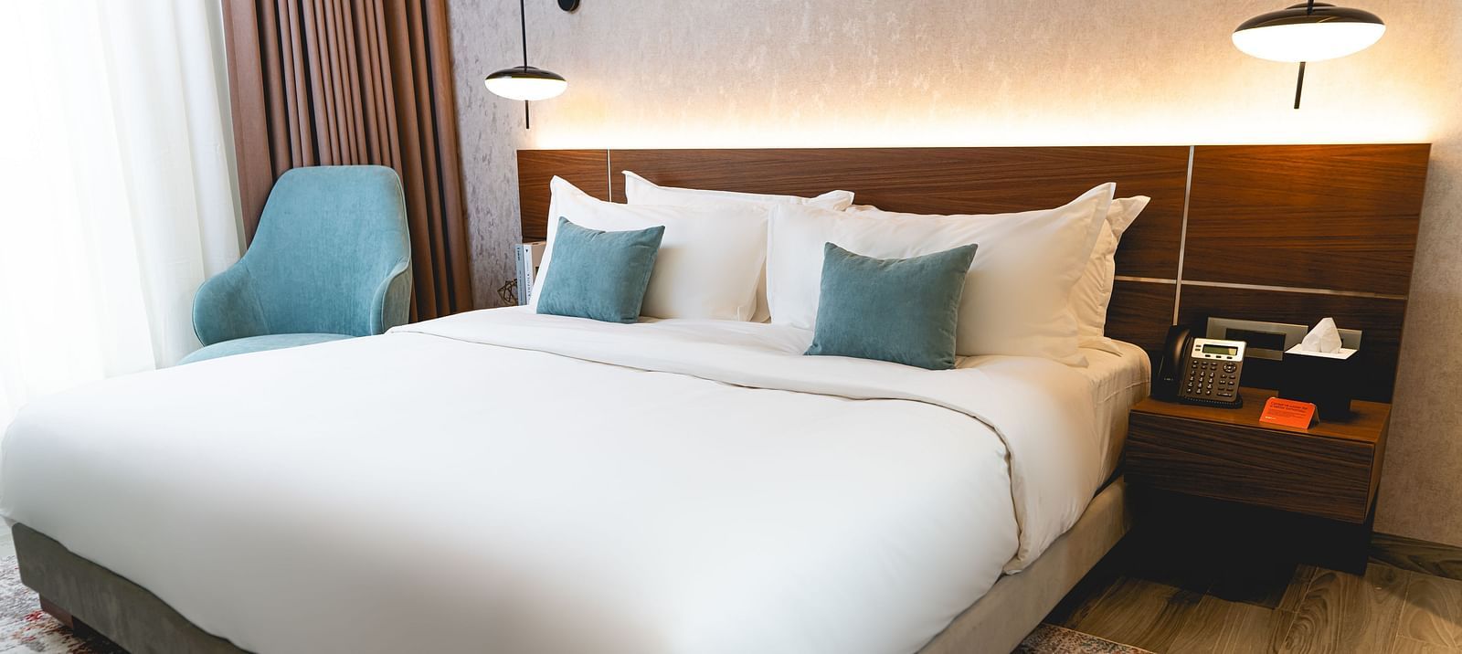 A bed and accent pillows at KertelSuites, featuring a wooden headboard, two bedside tables with lamps, and a armchair by the window
