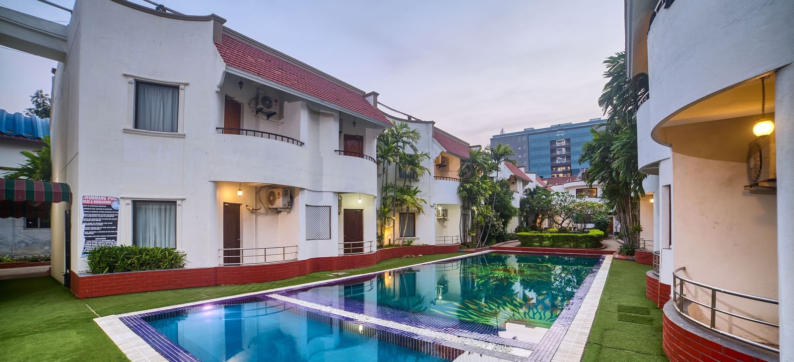Wide exterior view of the hotel building overlooking a sparkling blue swimming pool surrounded by green lawn at Ilara Hotels & Spa, OMR, Chennai.