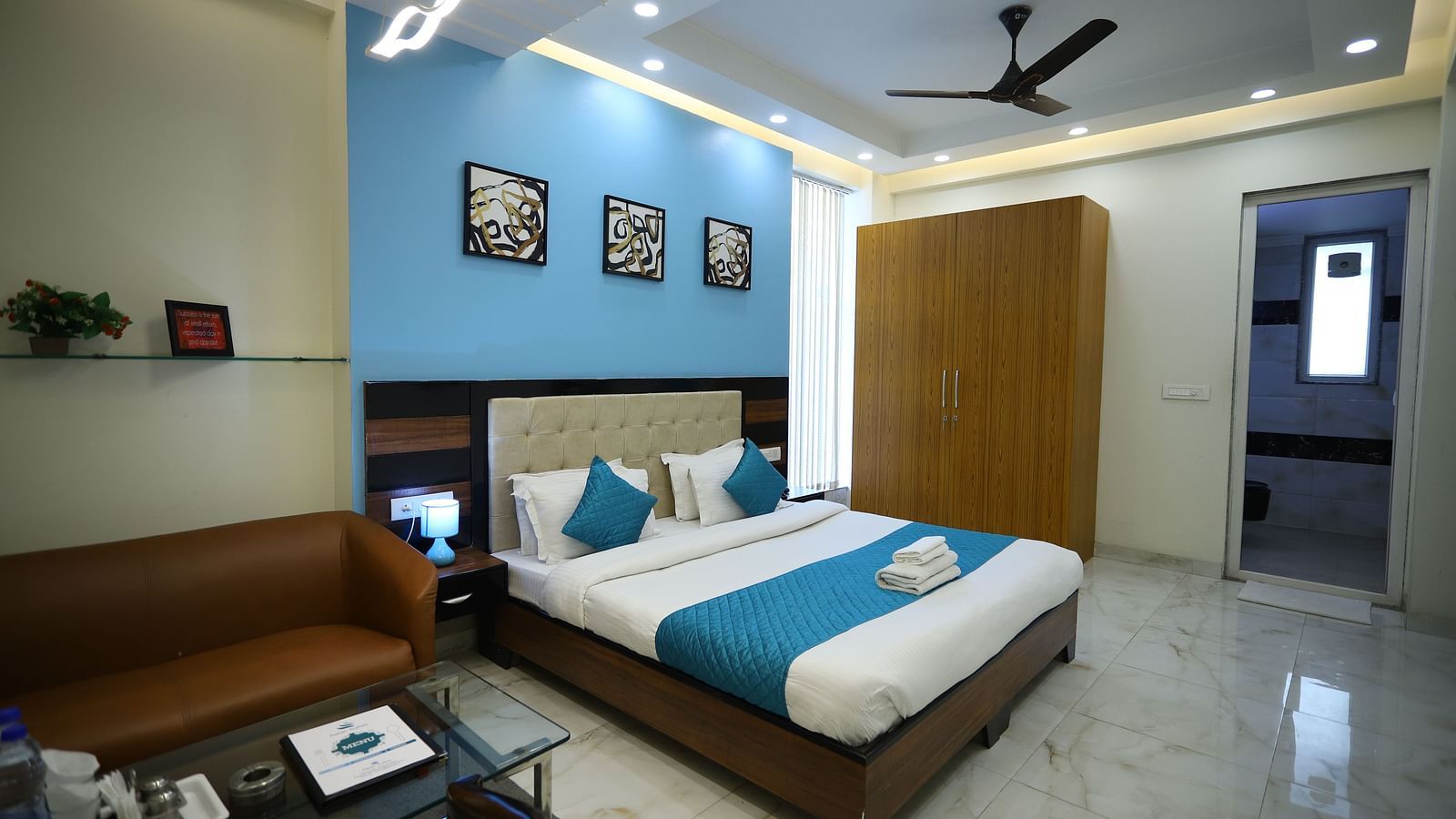 A room with a double bed, a sofa, a coffee table, a ceiling fan, and framed artwork on the wall - Asian Suites Cyber Park, Gurgaon