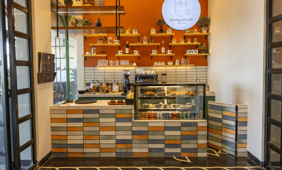 Cafe front at Hotel Bawa, Udaipur displaying a tiled counter, glass food cabinet, espresso machine, wall shelves, and circular overhead logo sign.