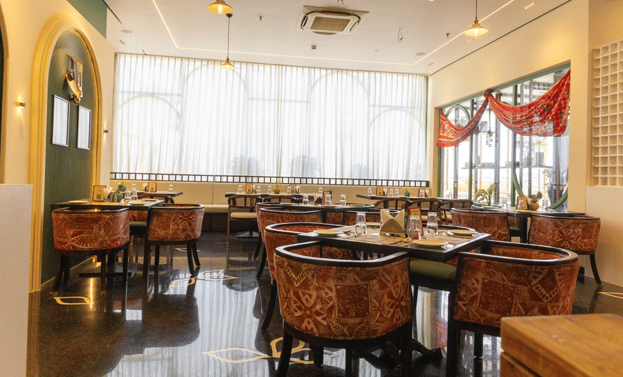 A restaurant at Hotel Bawa, Udaipur showing dining tables, patterned armchairs, sheer curtained windows, hanging lights, and polished flooring.