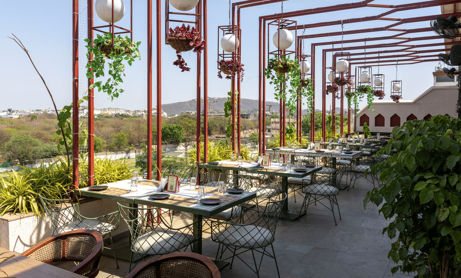 Outdoor rooftop dining section at Hotel Bawa, Udaipur showing metal canopy structures, hanging plant baskets, dining tables, and scenic views.