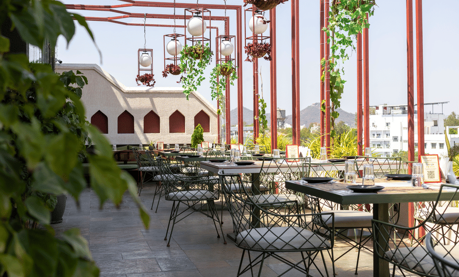 Outdoor rooftop terrace at Hotel Bawa, Udaipur featuring dining tables, chairs, metal overhead framework, hanging lantern lamps, and greenery.