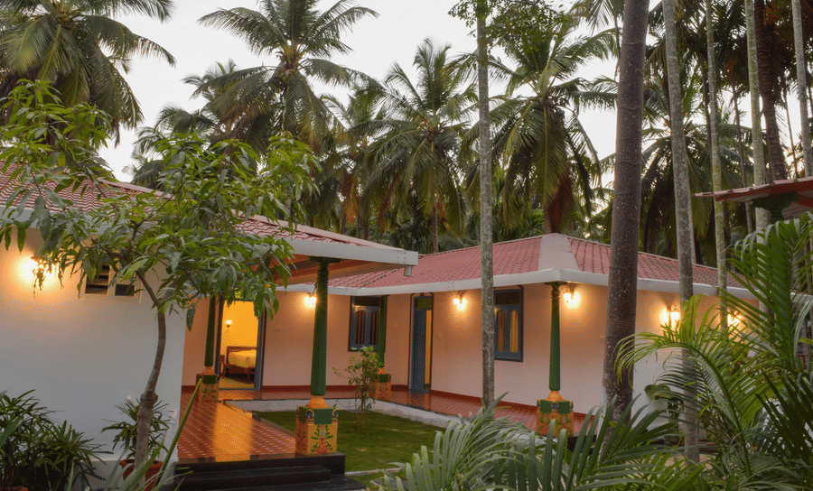 A white resort cottage with a red roof and blue window frames, surrounded by palm trees and green lawns - Ibex Resorts, Coimbatore (Kakarla)