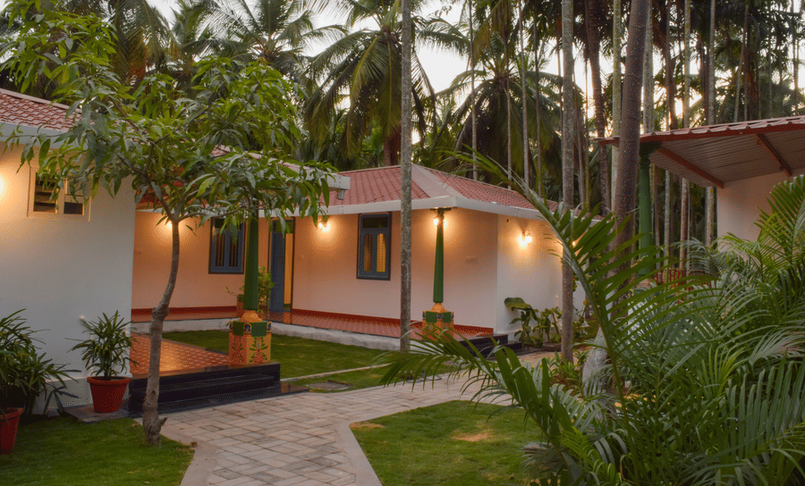 A white resort cottage with a red roof and blue window frames, surrounded by palm trees and green lawns - Ibex Resorts, Coimbatore (Kakarla)