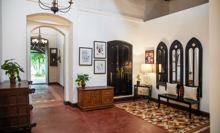 An image of a huge lobby where various decorative items are placed on top of the cabinets and shelves, there is another table with lamp and three vintage style window near it - Lamrin Ucassaim Hotel, Goa