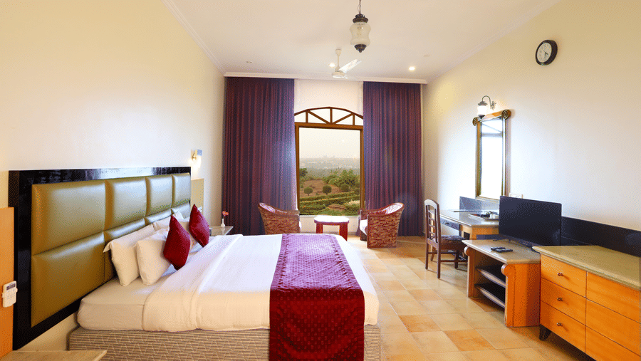 A bright hotel bedroom with a big bed, a desk and a TV, and 2 chairs by an arched window overlooking the greenery outside | Kohinoor Samudra Beach Resort, Ratnagiri