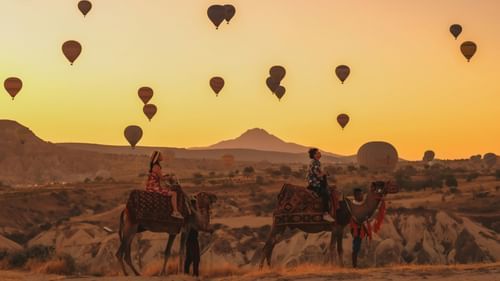 An image of hot air balloons in the air with a two women sitting on camels in the foreground and the orange sky during the sunset in the background