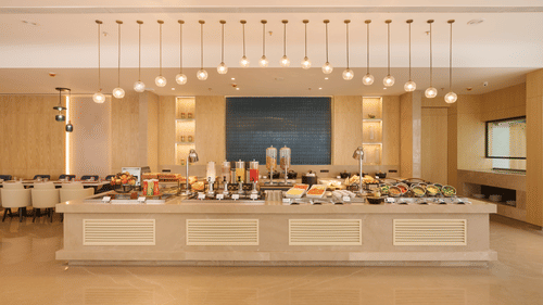 Skywings Sarovar Portico Mathura restaurant with wide buffet counter fully stocked, pendant lighting, blue wall art, and bright cream interiors.