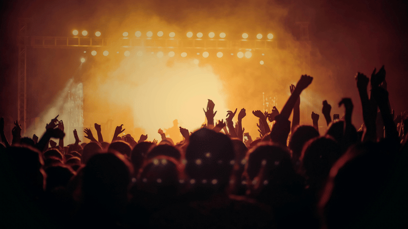 A blurred crowd silhouette with hands raised, facing an intense, hazy orange stage light at a concert.