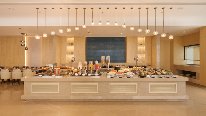 Skywings Sarovar Portico Mathura restaurant with wide buffet counter fully stocked, pendant lighting, blue wall art, and bright cream interiors.