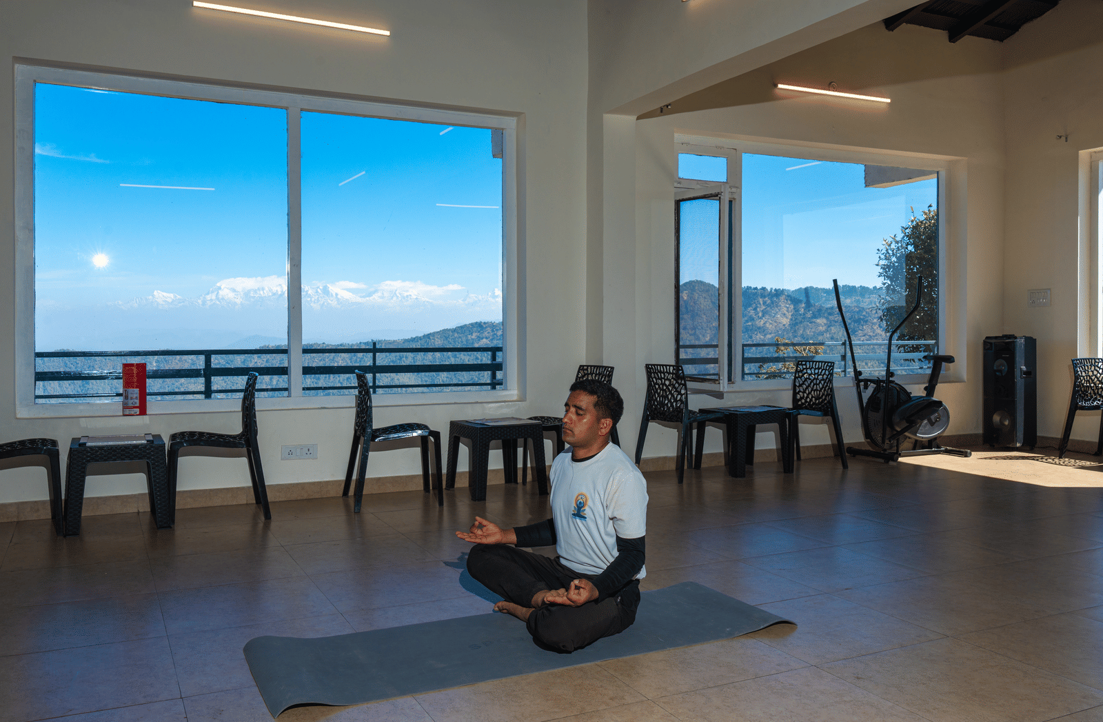 A person sitting in a meditative yoga pose on a mat in a bright studio with large windows at Himalayas Resort By The Lake Hill, Mukteshwar.
