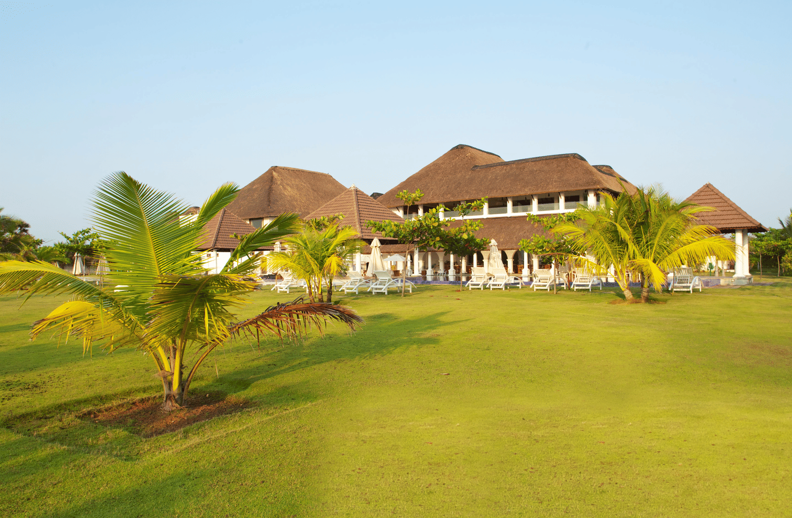 A wide green lawn with small palm trees situated in front of thatched-roof resort buildings | Le Pondy