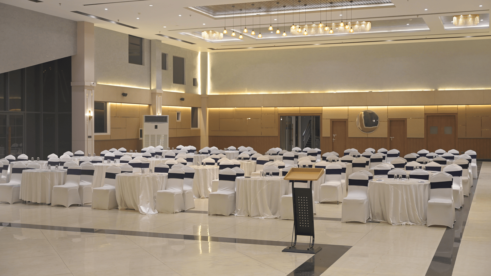 A front-angled view of the banquet hall set with round tables and white seating under warm ceiling lights at Hotel Sonar Bangla Mayapur.
