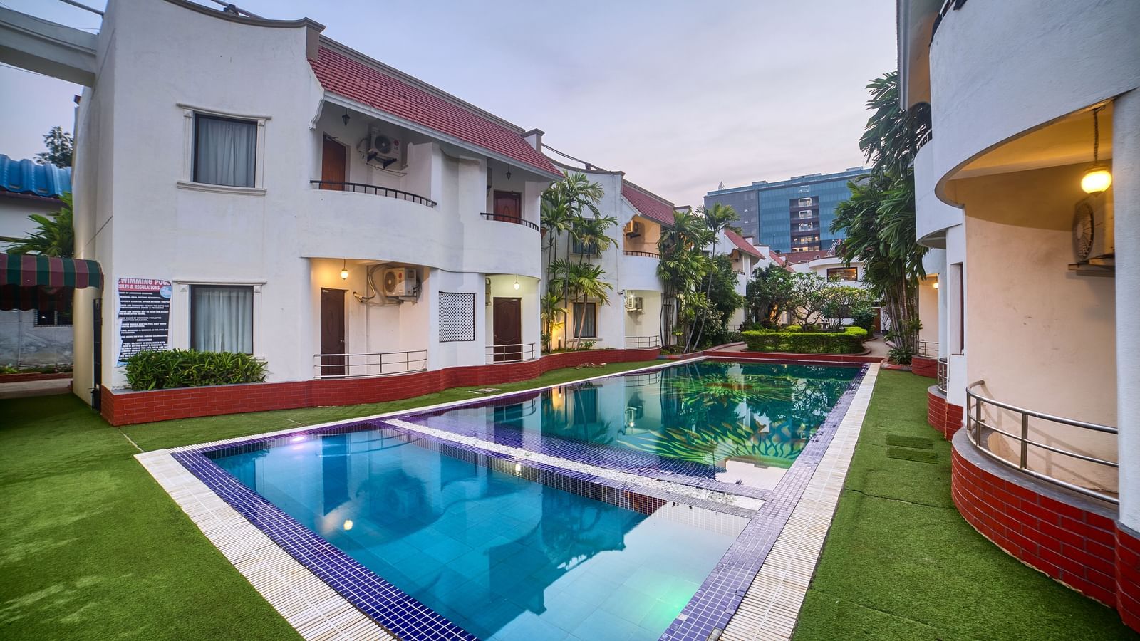 Wide exterior view of the hotel building overlooking a sparkling blue swimming pool surrounded by green lawn at Ilara Hotels & Spa, OMR, Chennai.