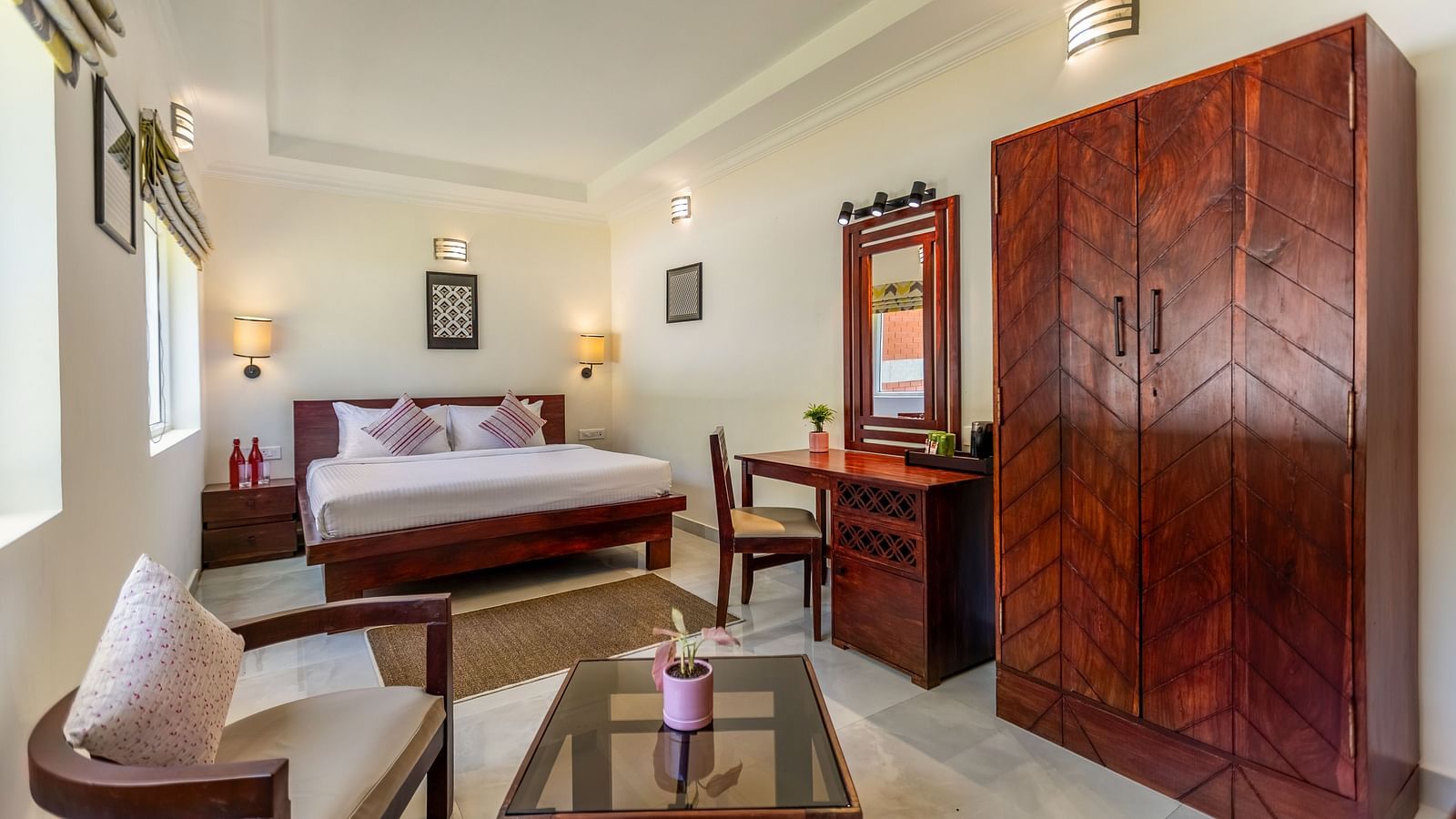 A bright, clean hotel room with a bed, a wooden desk and chair, a large wooden wardrobe, and a seating area with a glass-topped coffee table - Mango Hill Resort, Coimbatore