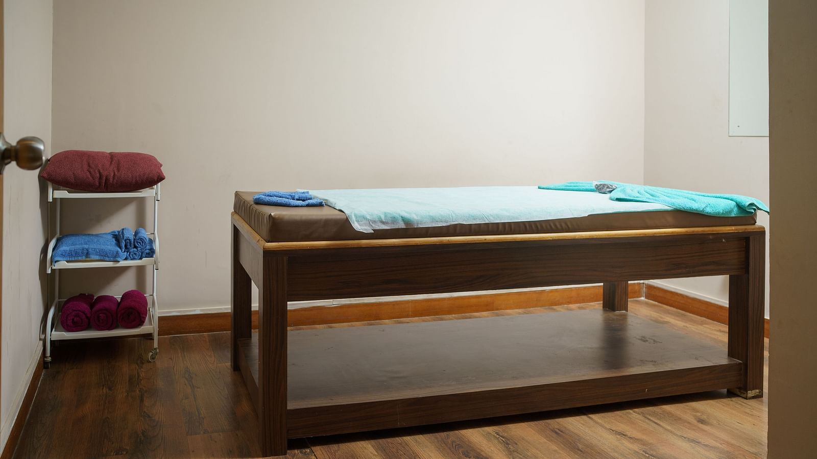 A wooden massage table in a room with light-coloured wooden flooring and white walls, with some folded towels on a shelf at Orsino Spa Resort.