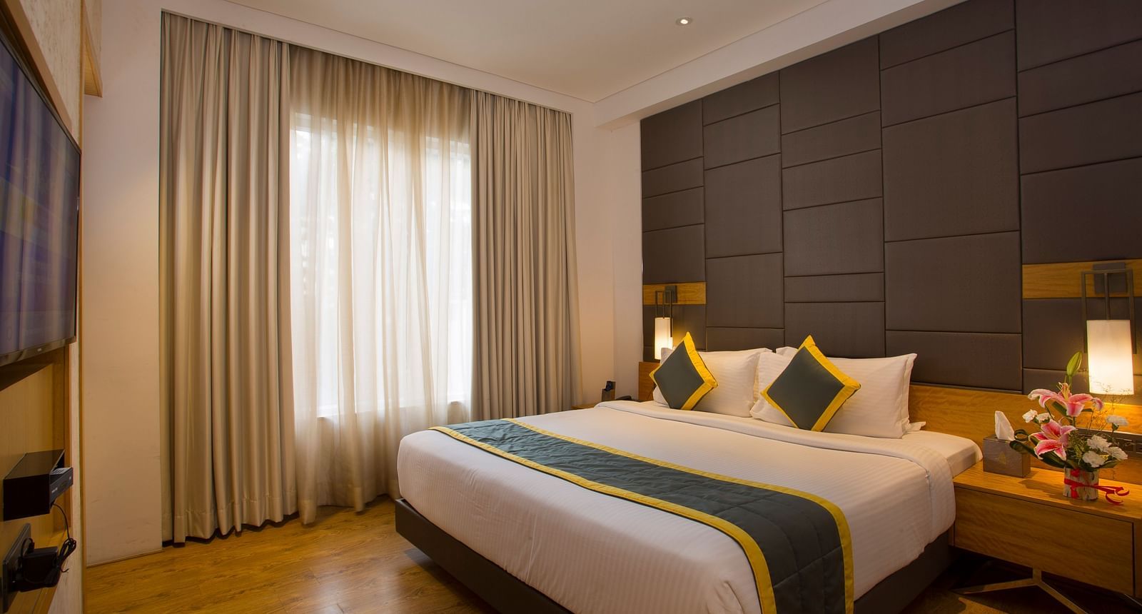 a bedroom with a large bed opposite a television mounted on the wall and side drawers on each side - Hotel Fountain Tree By TGI - JP Nagar, Bangalore