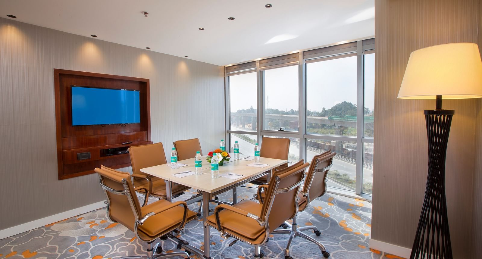 A small meeting room with a round table, tan leather chairs and a TV, with a large window and a floor lamp - Hotel Southend by TGI - Bommasandra, Bangalore