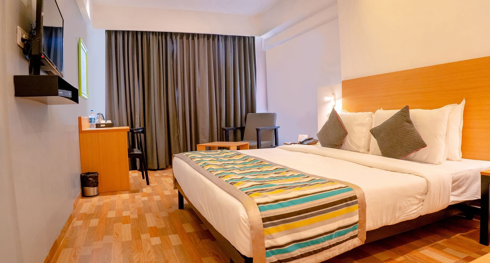 Another view of a cosy hotel room with a large bed, colourful throw and wooden furniture, including a desk with a TV above it - TGI Ameya Grand, Goa