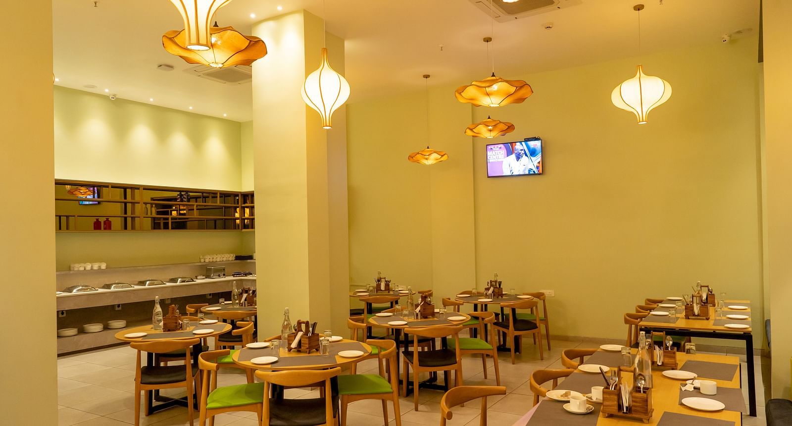 Another angle of the restaurant interior showing tables set for dining with hanging orange pendant lights and a TV screen on the wall - TGI Ameya Grand, Goa