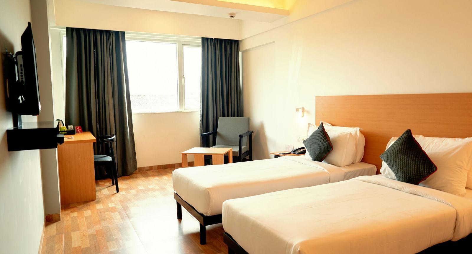 A bright hotel room with two single beds, a desk and a flat-screen TV, with a large window draped in gray curtains - TGI Ameya Grand, Goa