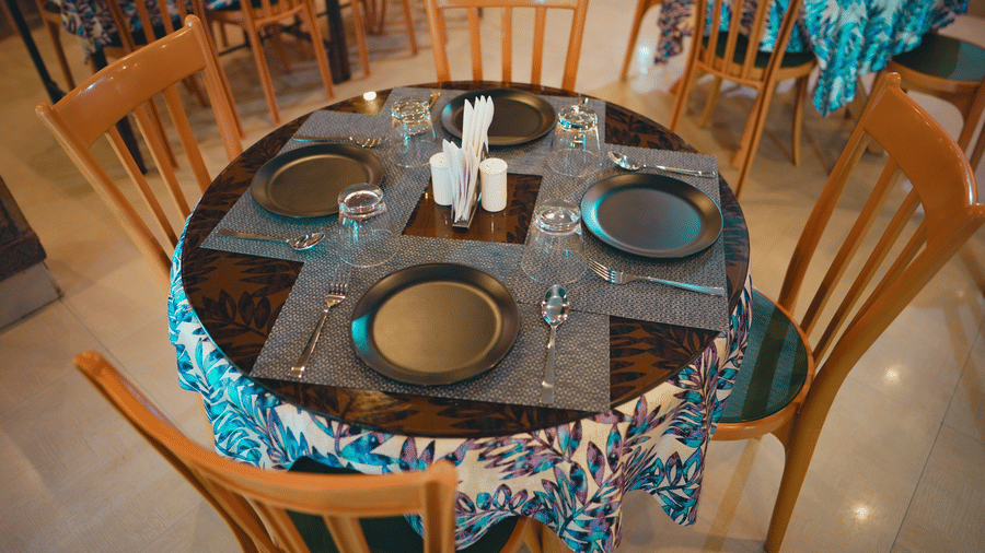 A set-up round restaurant dining table with wooden chairs, a patterned tablecloth, and formal place settings.