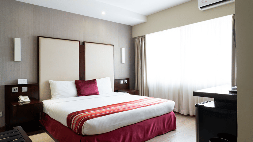 A hotel room at The Zehneria, Nairobi, featuring a double bed, an AC unit, a writing table, and night stands on either sides of the bed.