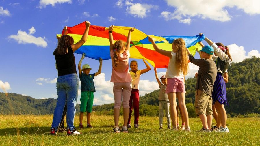  A group of diverse children and adults are playing a game with a colorful parachute in a grassy field on a sunny day.