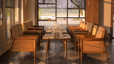 An indoor seating area with wooden chairs and large windows framing outdoor garden views at Hotel Sonar Bangla, Mayapur.