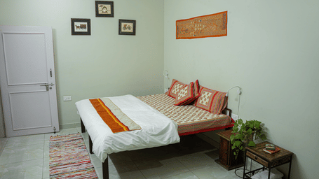A Deluxe Room With Balcony at Jaypore360 Homestay featuring a neatly-made bed with a metal frame, a nightstand, a potted plant for decoration, and framed paintings on a wall.