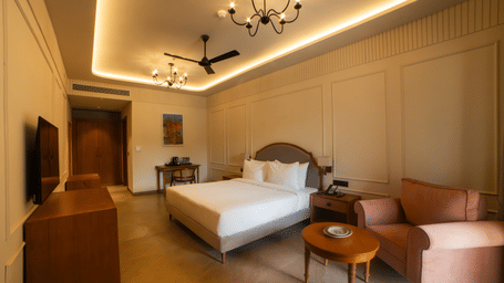 A room with a king size bed, a pink armchair with a small round table, wooden furniture, and a chandelier under a recessed lighted ceiling at Jivana Wellness Retreat, Bhopal.