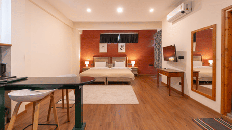 A wide-angle view of a studio room featuring wooden flooring, a small kitchenette, and twin beds against an orange accent wall - Johnson Lodge & Spa, Manali
