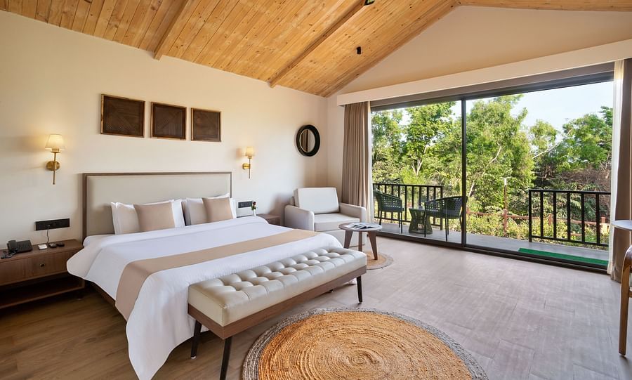 An image of a premium cottage featuring a king size bed, a bedside table and lamp on either side, a sofa chair, and a spacious balcony attached to the room at Stone Woods Premier Resort, Gokarna