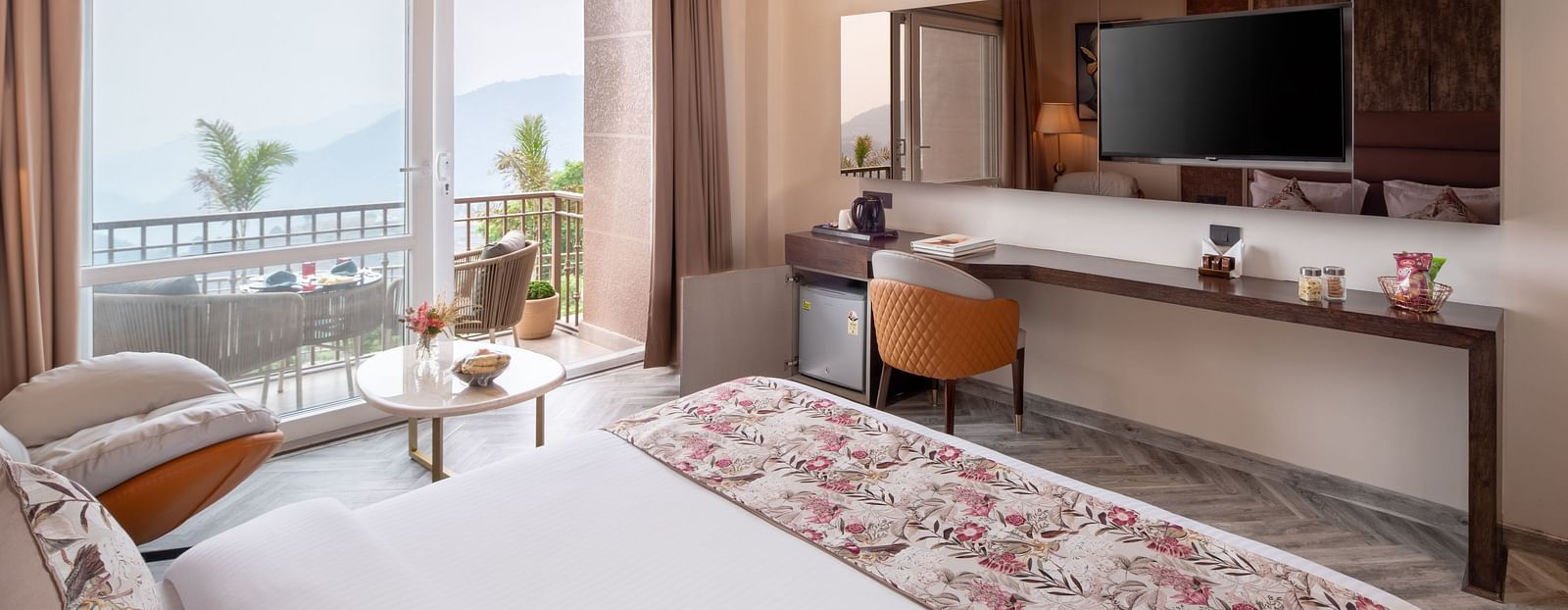 an image of a bedroom with balcony from where sunlight is flooding the room which has a flat TV screen,  a narrow desk below it paired with a chair.