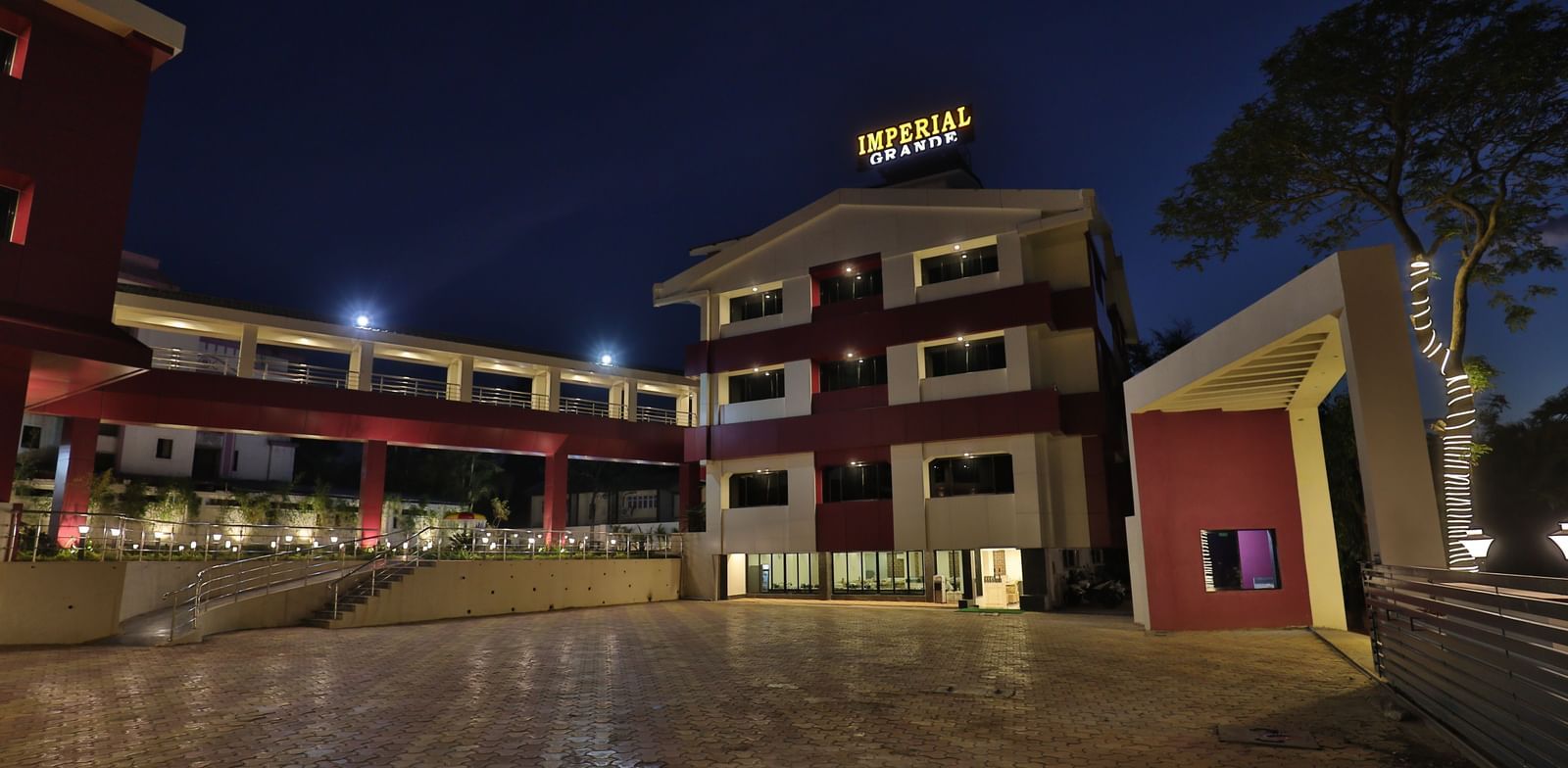The exterior of Imperial Grande at night, showing a multi-story building with illuminated logo and a paved courtyard.
