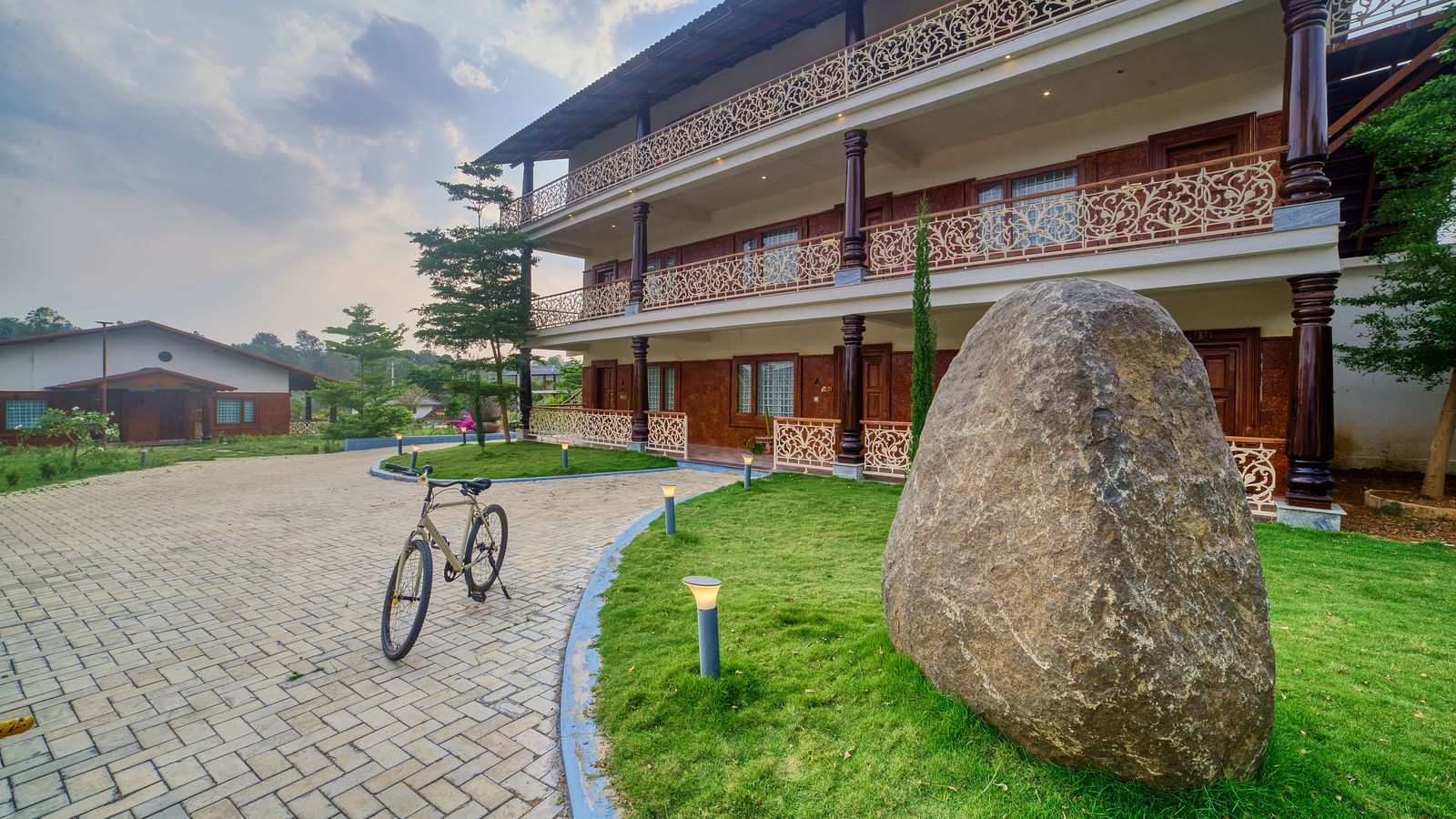 The exterior of a multi-storey building, possibly a resort or hotel, with a pathway leading up to it, flanked by a large rock and some greenery - Vintage Trails by Ilara