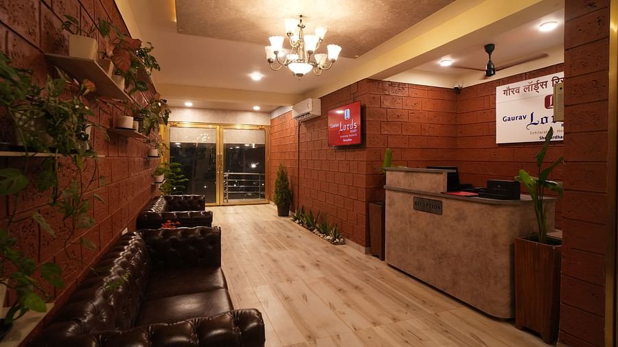 Interior hallway of a resort featuring a reception desk, dark leather sofas, potted plants on a brick wall, and a chandelier - Gaurav Lords Resort Shrivardhan.