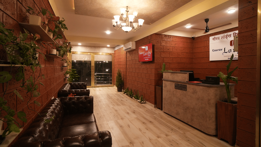Interior hallway of a resort featuring a reception desk, dark leather sofas, potted plants on a brick wall, and a chandelier - Gaurav Lords Resort Shrivardhan.