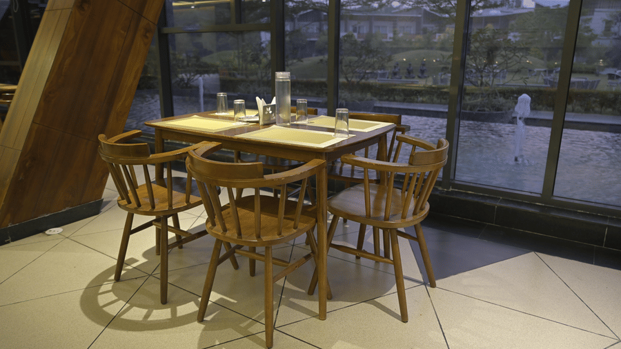 A wooden dining table with 4 wooden chairs next to a glass window at Hotel Sonar Bangla, Mayapur.
