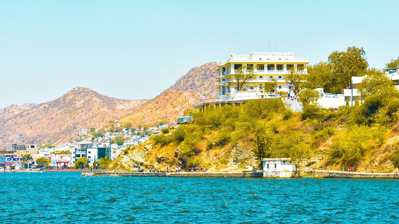 A historic fort stands overlooking by calm water and lush green bushes.