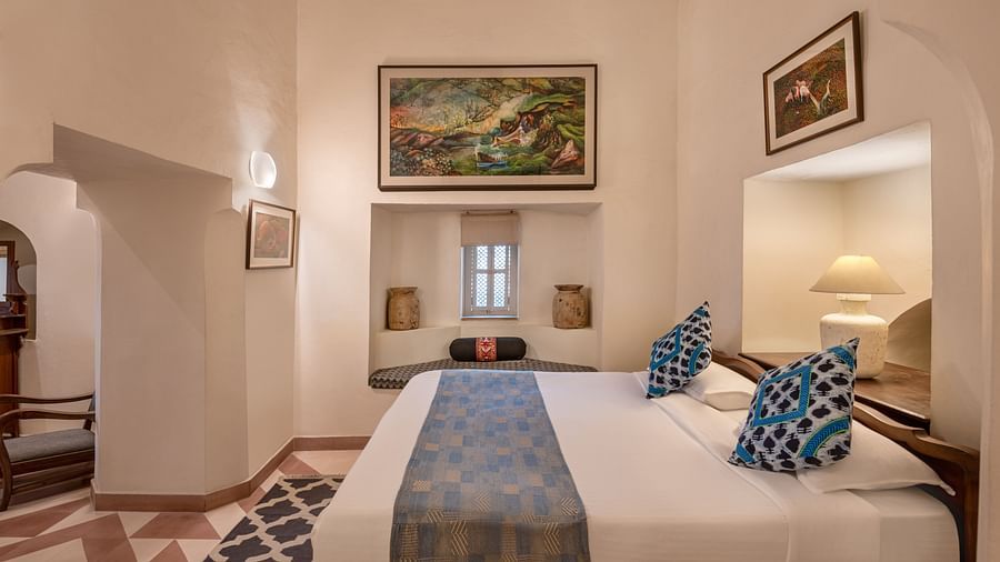 A room with a bed, two framed artworks, a window seat, and a small window in Tijara Fort-Palace - 19th Century, Alwar.