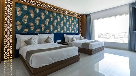 A bedroom at the Bamboo Saa Mulberry Resort featuring two twin beds with white linens and grey throws. A decorative wall panel with a blue and gold pattern is above the beds and a large windows is visible.